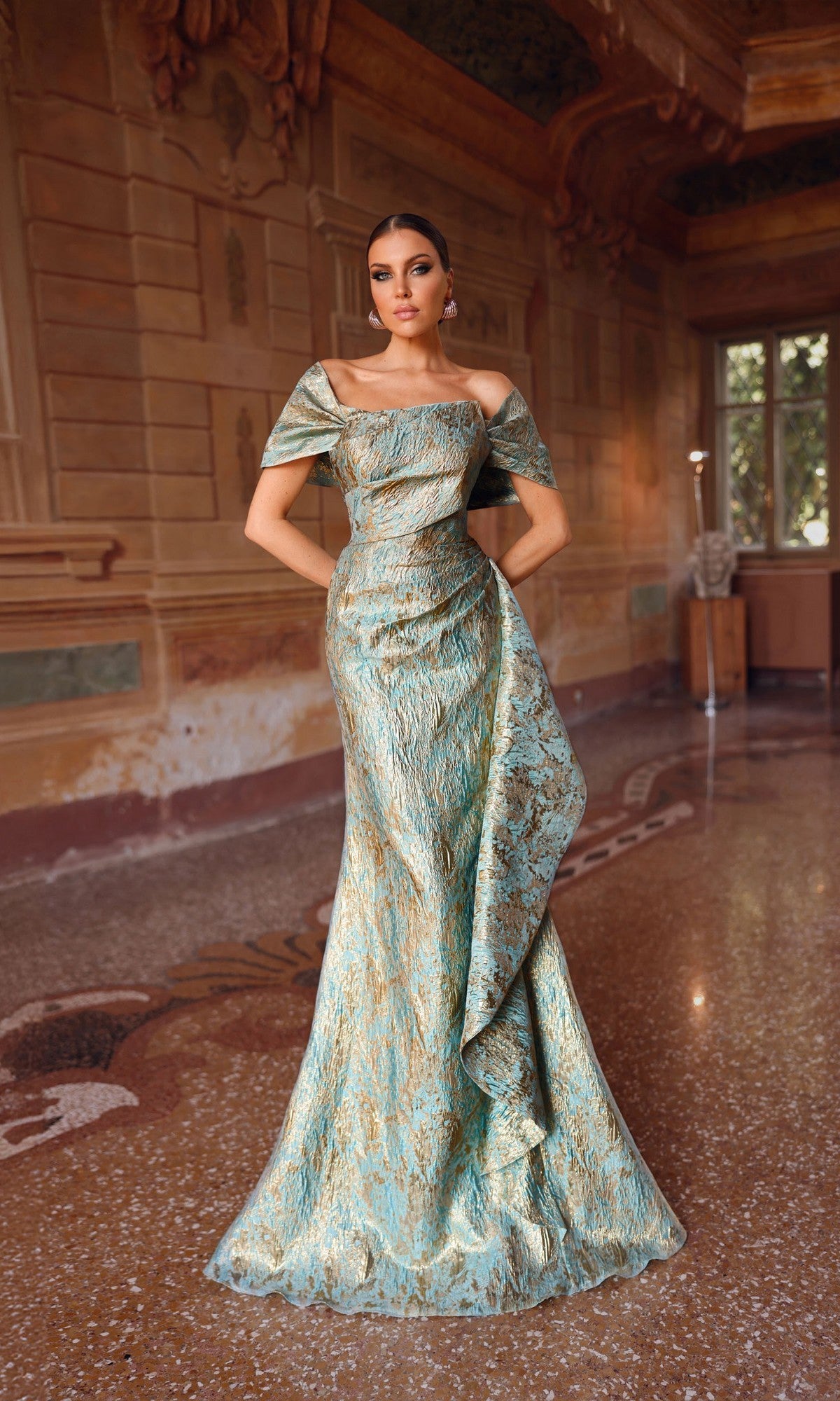A woman stands indoors in the Alyce 27750 long formal dress—a metallic jacquard brocade, off-the-shoulder mermaid gown—set against ornate walls and a polished stone floor that enhance her elegant look.